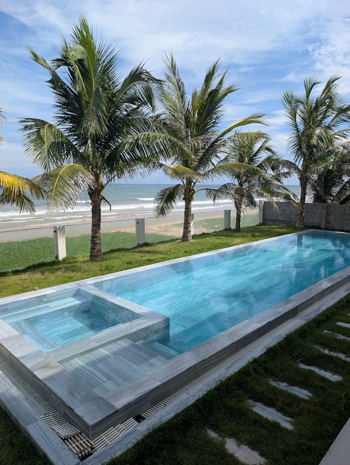 Stunning coastal view with a modern pool and palm trees beside a sandy beach.